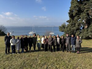 Group photo of 17 smiling workshop attendees. Behind them is a lake and a large tree.